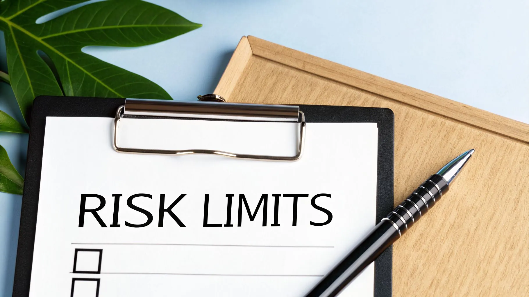 A clipboard with a paper showing 'RISK LIMITS' and empty checkboxes, next to a pen and a plant.